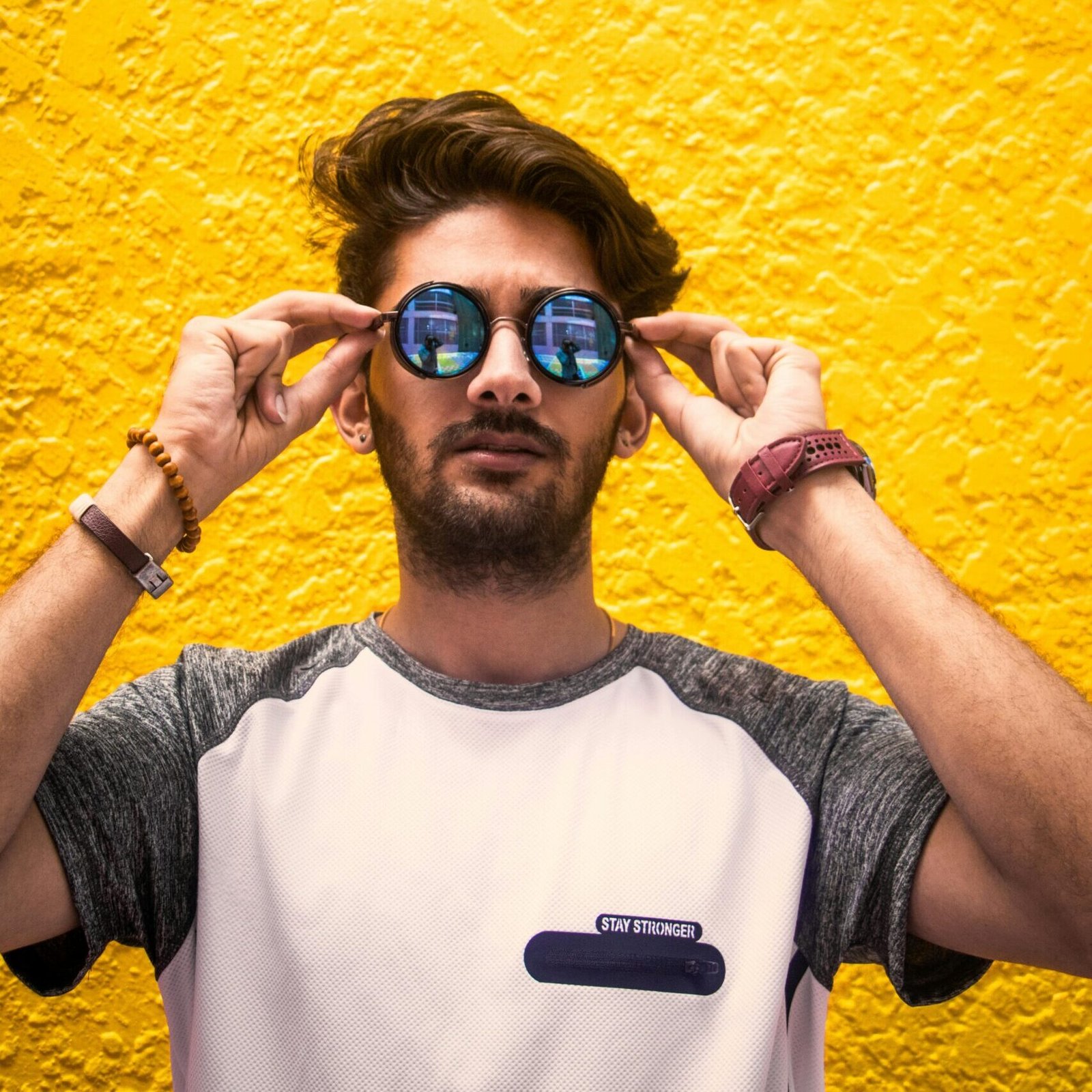 Young man with sunglasses standing against a vibrant yellow wall in daylight.