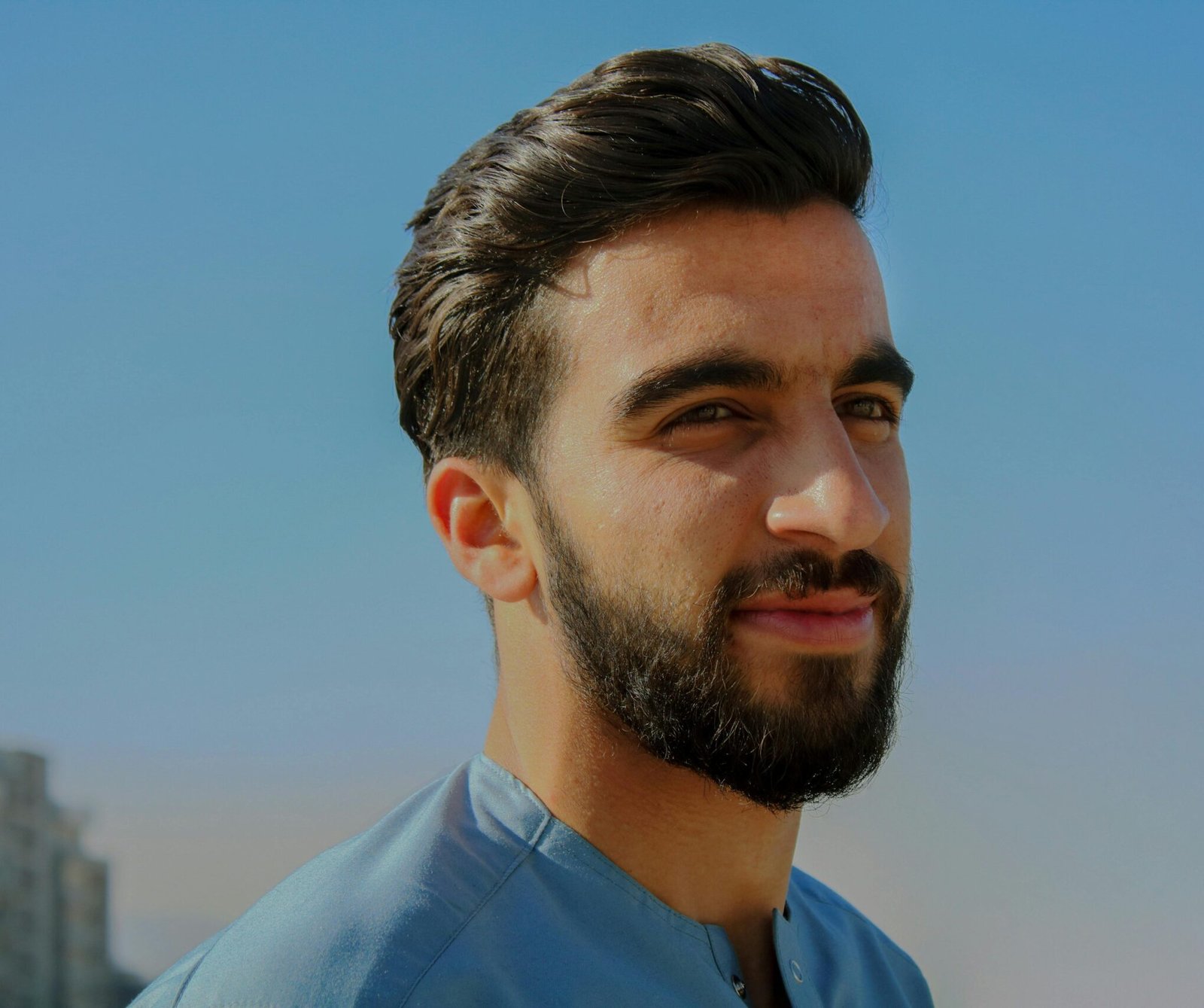 A close-up outdoor portrait of a young man with facial hair, exuding confidence under a clear sky.